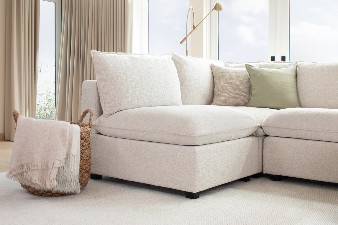 A modern living room featuring the Kova L Shape with Ottoman, adorned with a light green throw pillow. A basket with folded white and grey blankets sits nearby. Large windows with beige curtains let in natural light, creating a cozy and inviting atmosphere.