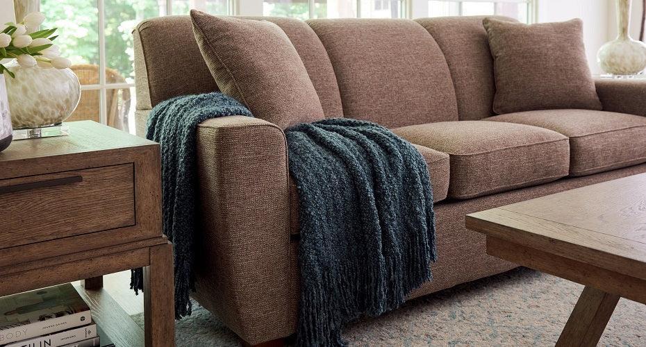A cozy living room featuring a brown sofa adorned with a dark blue fringed throw blanket draped over the armrest. The room includes a wooden side table with a floral arrangement and a view of greenery through a large window in the background.