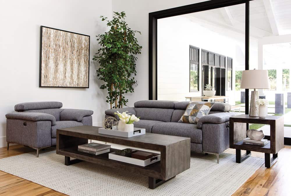 A modern living room featuring a gray sofa and armchairs, a rectangular wooden coffee table with storage, and a tall green plant. Large sliding glass doors open to an outdoor patio. A framed abstract artwork hangs on a white wall. The room has a cozy ambiance.