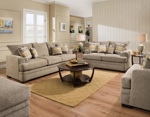 A cozy living room featuring two beige sofas with matching plaid pillows, a round wooden coffee table, a beige area rug, and decorative wall art. The room also has wooden flooring, a window with white trim, and a lamp on an end table near the corner.