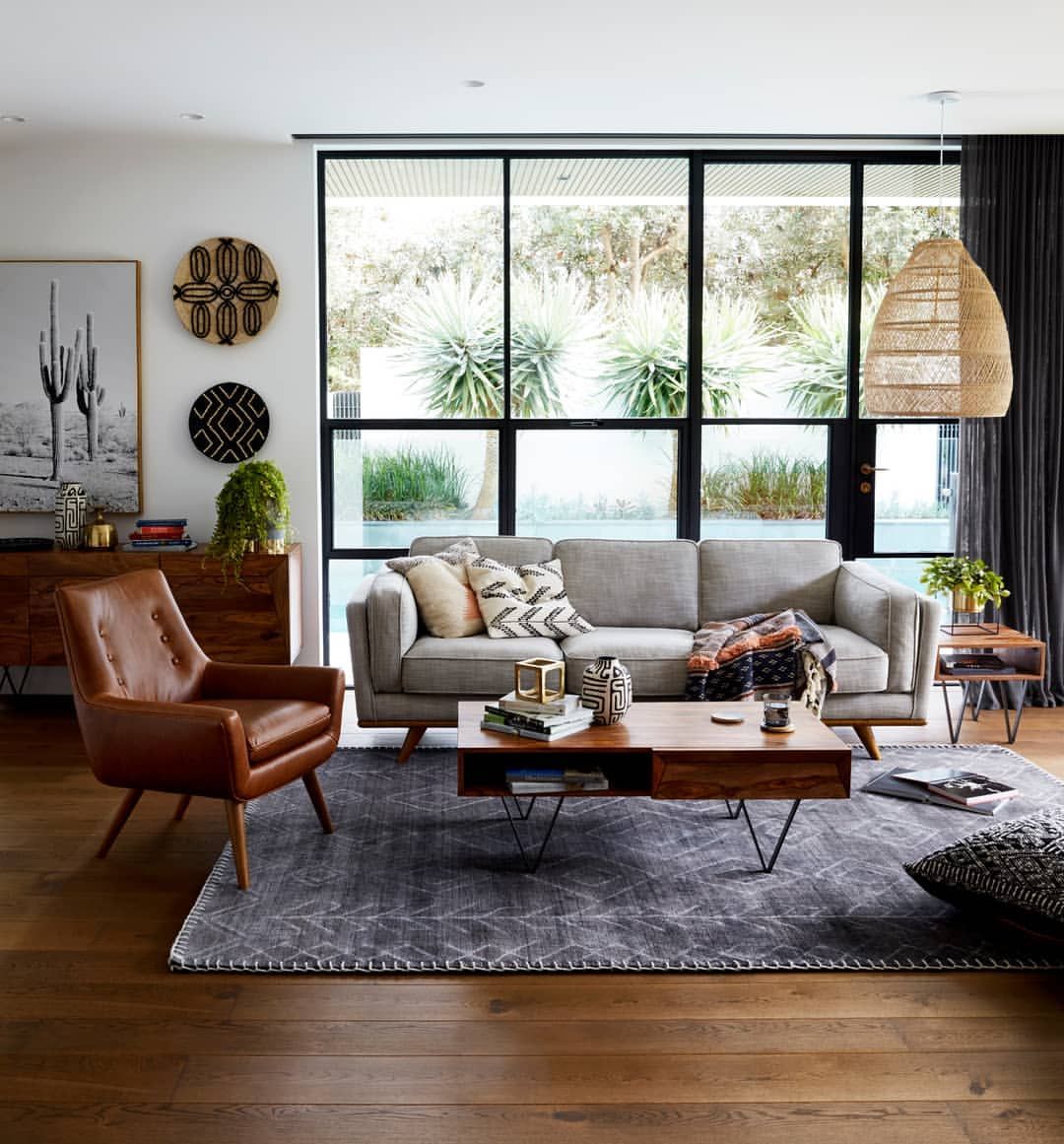A modern living room with a large window showing a garden view. It features a gray sofa, brown leather armchair, wooden coffee table, and patterned rug. Decor includes a wall art of a cactus, woven baskets, a wooden sideboard, and a pendant light.