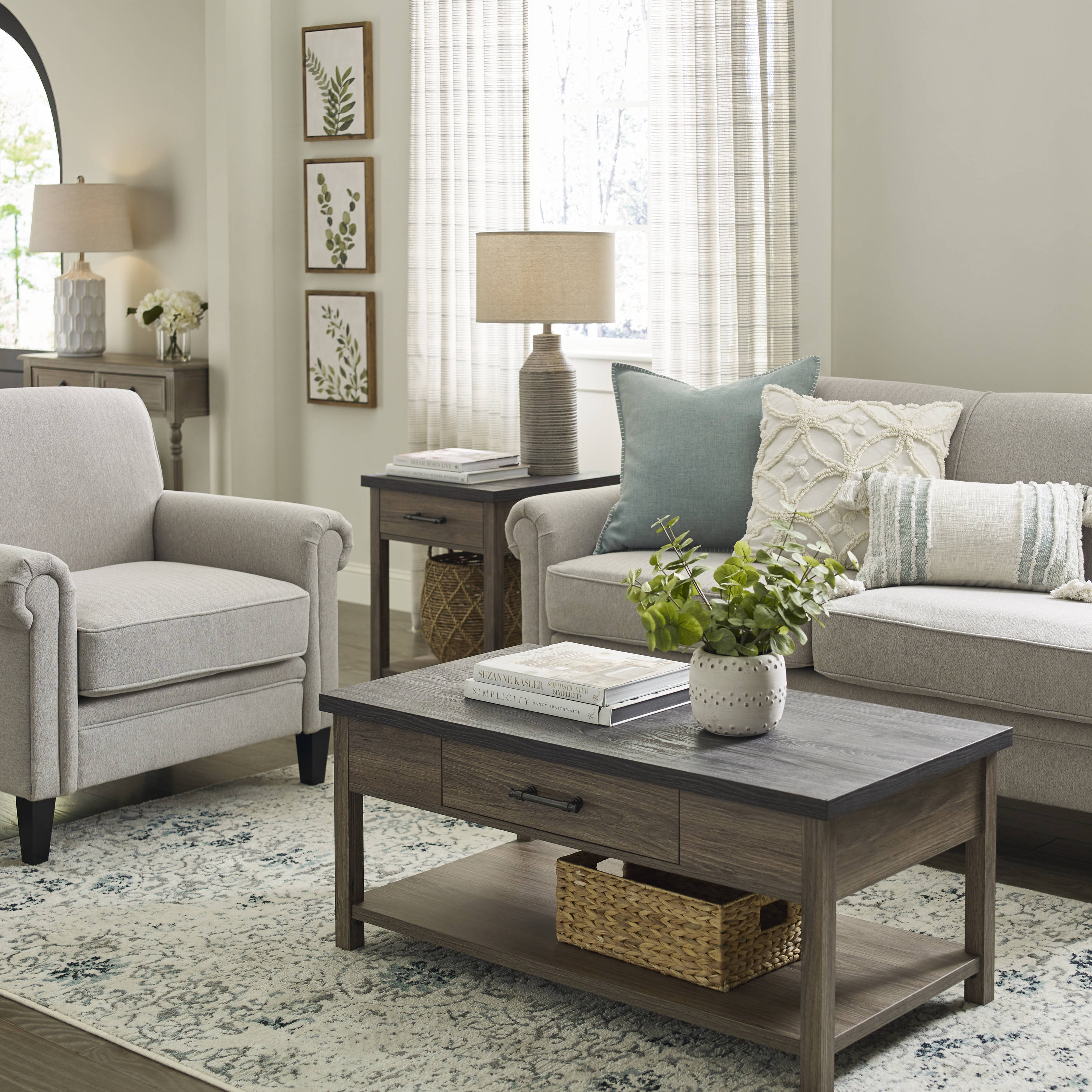 A cozy living room features a beige armchair, a light gray sofa with patterned pillows, a wooden coffee table with a drawer and shelf holding a woven basket and a plant, a side table with a lamp, and three botanical prints on the wall. Sunlight filters through sheer curtains.