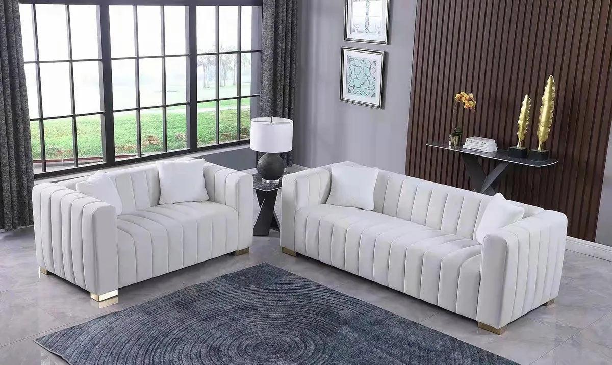 A modern living room with two white sofas facing each other, each adorned with white pillows. A small black lamp sits on a table between the sofas. A large window with curtains allows natural light in. A rug and decorative pieces complete the sleek design.