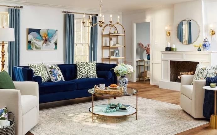 A cozy and elegant living room with a navy-blue velvet sofa adorned with patterned accent pillows. A glass coffee table with floral décor sits atop a light rug. The room features gold accents, a white fireplace, bookshelves with decor items, and large windows with teal curtains.