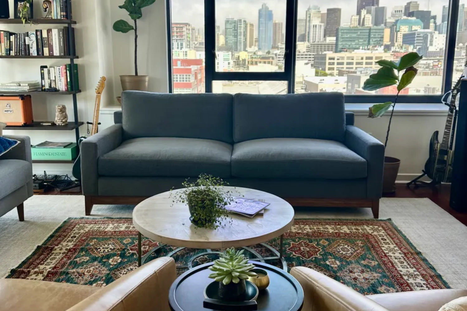 A modern living room with a gray sofa, a round wooden coffee table, and a potted plant on it. A vibrant patterned rug lies underneath. Large windows show a cityscape view. Books and a guitar are visible in the background, with a tall plant to the side.