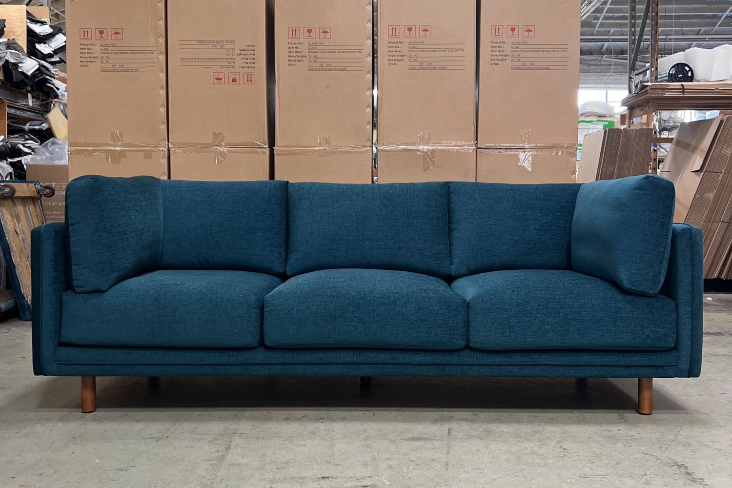 A modern teal sofa with plush cushions and wooden legs is displayed in a warehouse setting. Cardboard boxes are stacked in the background, and the surroundings suggest an industrial or storage space.