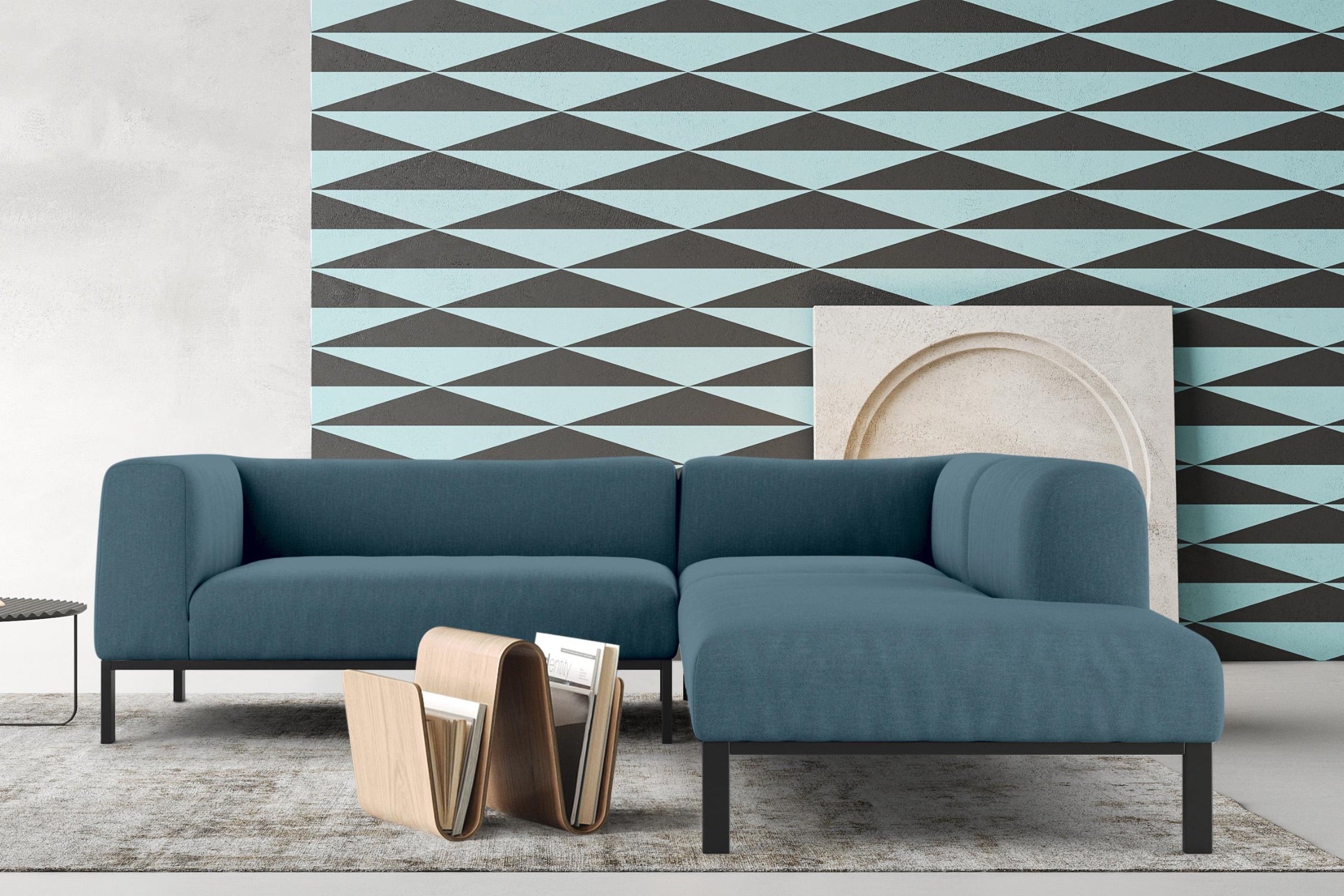 A modern living room featuring a blue L-shaped sofa on a muted rug. Behind the sofa is a geometric wallpaper with blue and black diamond shapes. A small side table and a wooden magazine rack complete the contemporary decor.