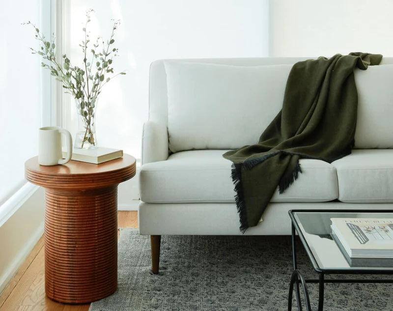 A minimalist living room showcases a white sofa adorned with a dark green throw, positioned next to a round wooden side table with a white mug, book, and a vase with foliage. A glass coffee table sits nearby on a patterned gray rug. Bright natural light fills the space.
