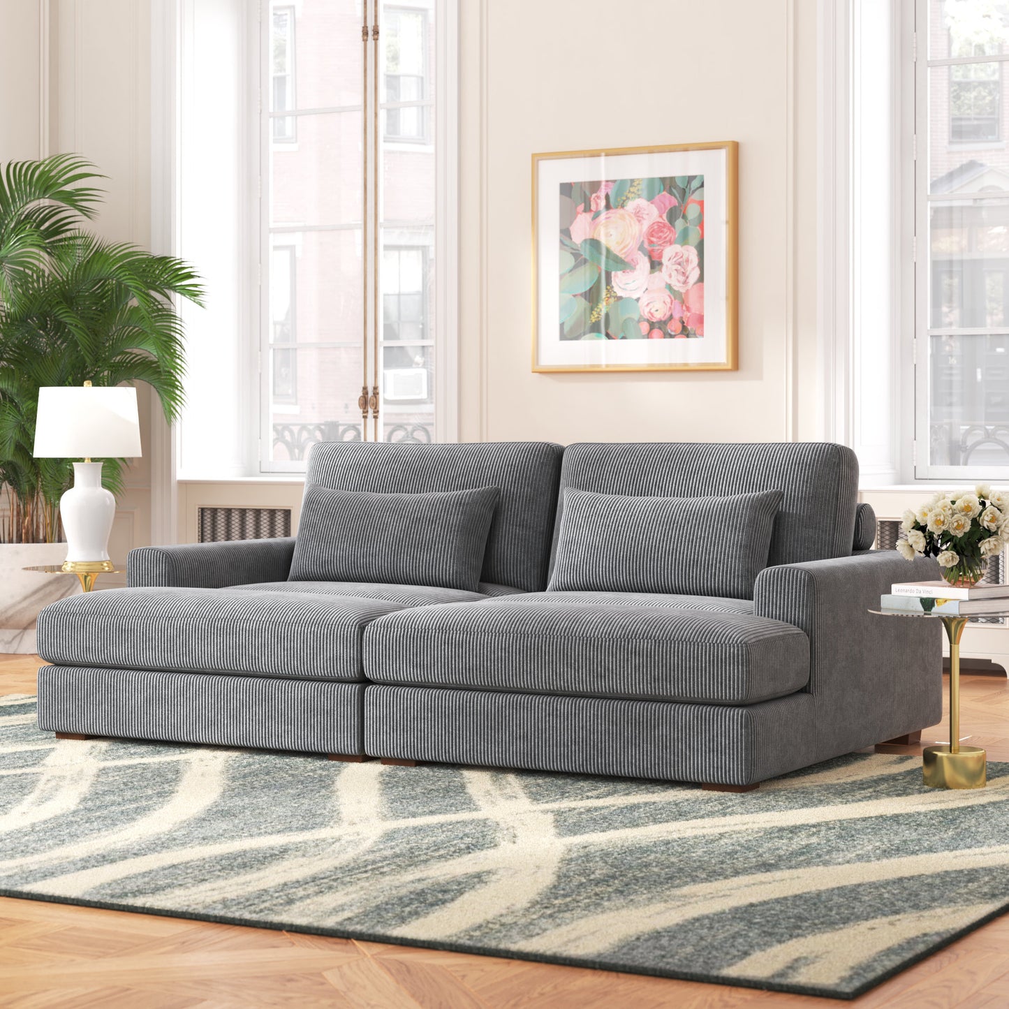 A cozy living room featuring the Etta Avenue Mordecai 90'' Square Arm Sofa in gray, complemented by a striped rug and a small side table with a lamp. A large plant is positioned beside the window, while a framed floral painting decorates the wall, illuminated by natural light streaming through tall windows.