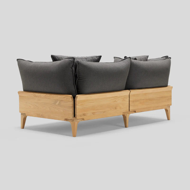 The Bluff Loveseat is a wooden sofa with a light finish, showcasing dark gray, plush back cushions. It boasts a modern design and angled wooden legs, set against a plain gray background.