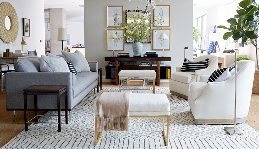 A modern living room featuring a gray sofa, two white armchairs with black and white striped cushions, and two cushioned ottomans. A wooden console table with decorative items, plants, and framed art is against the wall, and a large patterned rug covers the floor.