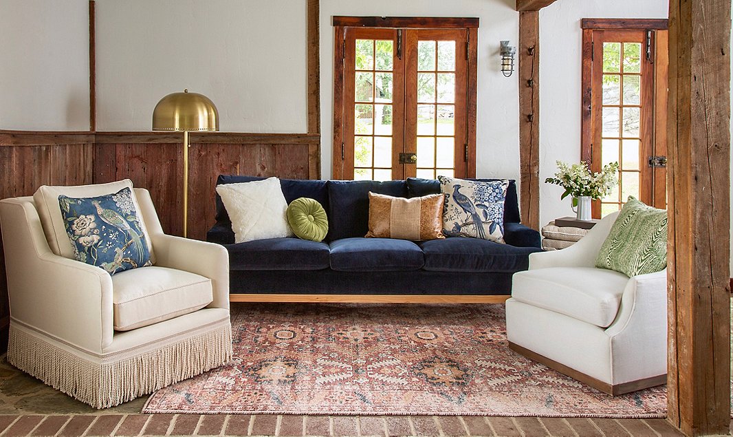 A cozy living room with a dark blue velvet sofa adorned with various cushions, flanked by two white armchairs. One armchair has fringe at the bottom. A vintage area rug, wooden beams, and a brass floor lamp complement the setting, with French doors in the background.