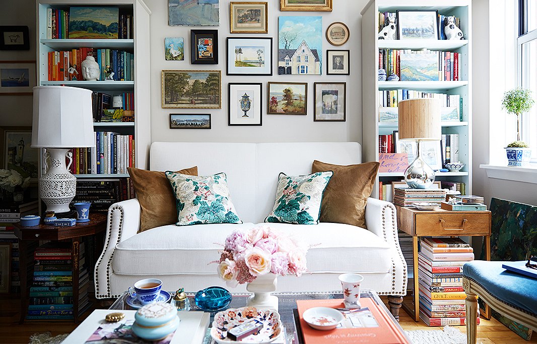 A cozy living room with a white sofa adorned with floral and brown throw pillows. The walls feature an eclectic gallery of framed art. Bookshelves filled with colorful books flank the sofa. A coffee table holds flowers, decorative trays, and assorted decor items.