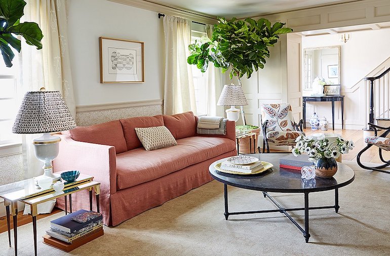 A cozy living room featuring a salmon-colored sofa with two patterned cushions, flanked by two side tables with lamps. A black coffee table with flowers and books sits on a beige carpet. Large green plants add a touch of nature, and a framed artwork decorates the wall.