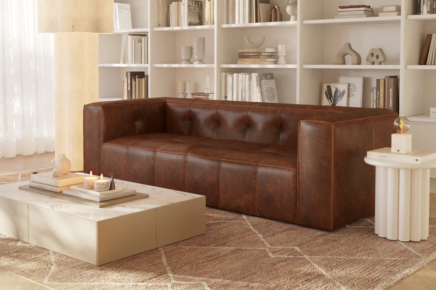 A cozy living room with a brown leather sofa in front of a marble coffee table. The table holds lit candles and books. Behind the sofa, white bookshelves display books and decor. Neutral tones and natural light enhance the serene atmosphere.