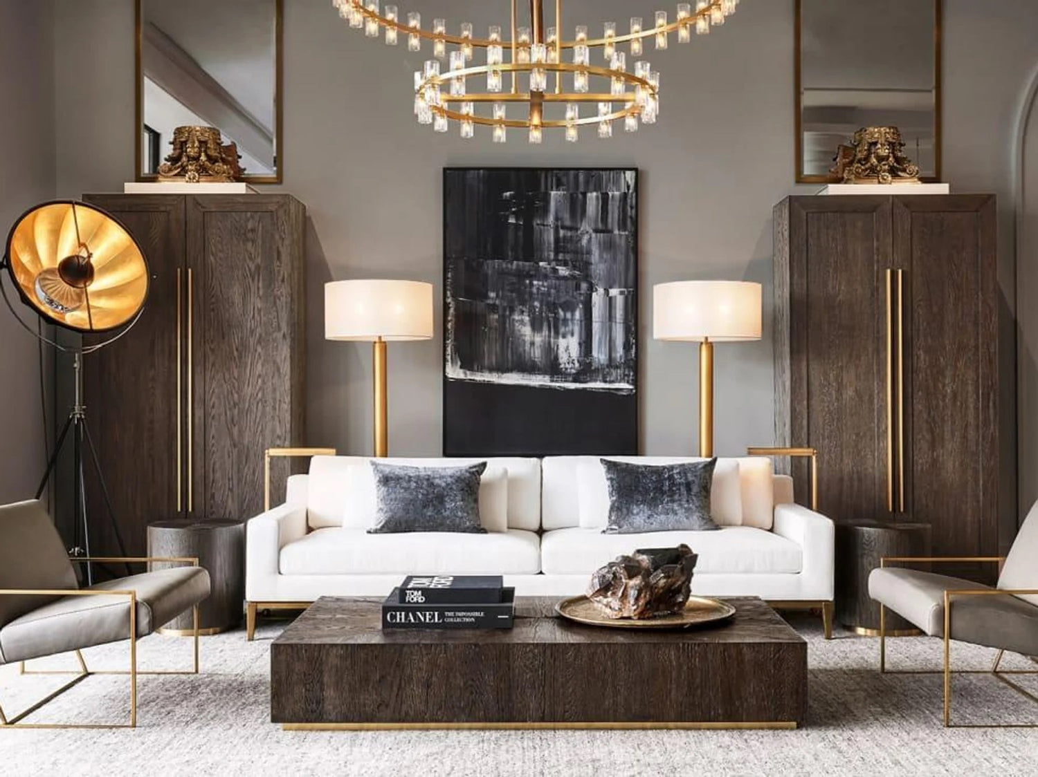 Stylish living room with a white sofa, dark wooden coffee table, and two modern armchairs. The room features a large chandelier, two floor lamps, a black and white abstract painting, and two wooden cabinets. A decorative piece and books are on the coffee table.