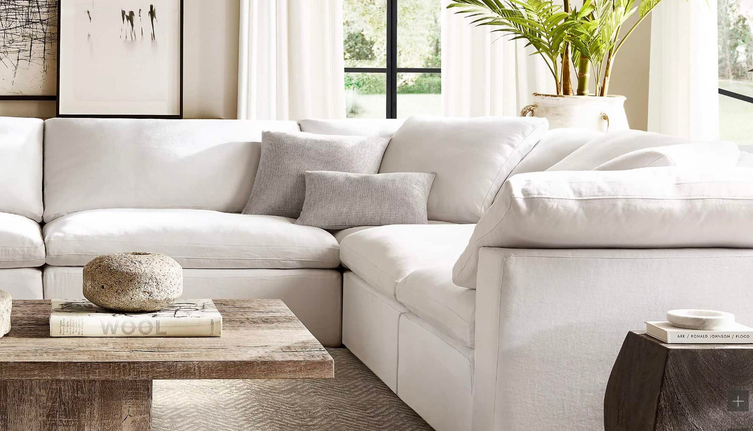 A bright and airy living room featuring a white sectional sofa with a grey accent pillow, a rustic wooden coffee table adorned with books and a stone sculpture, and a potted plant beside a large window with white curtains.