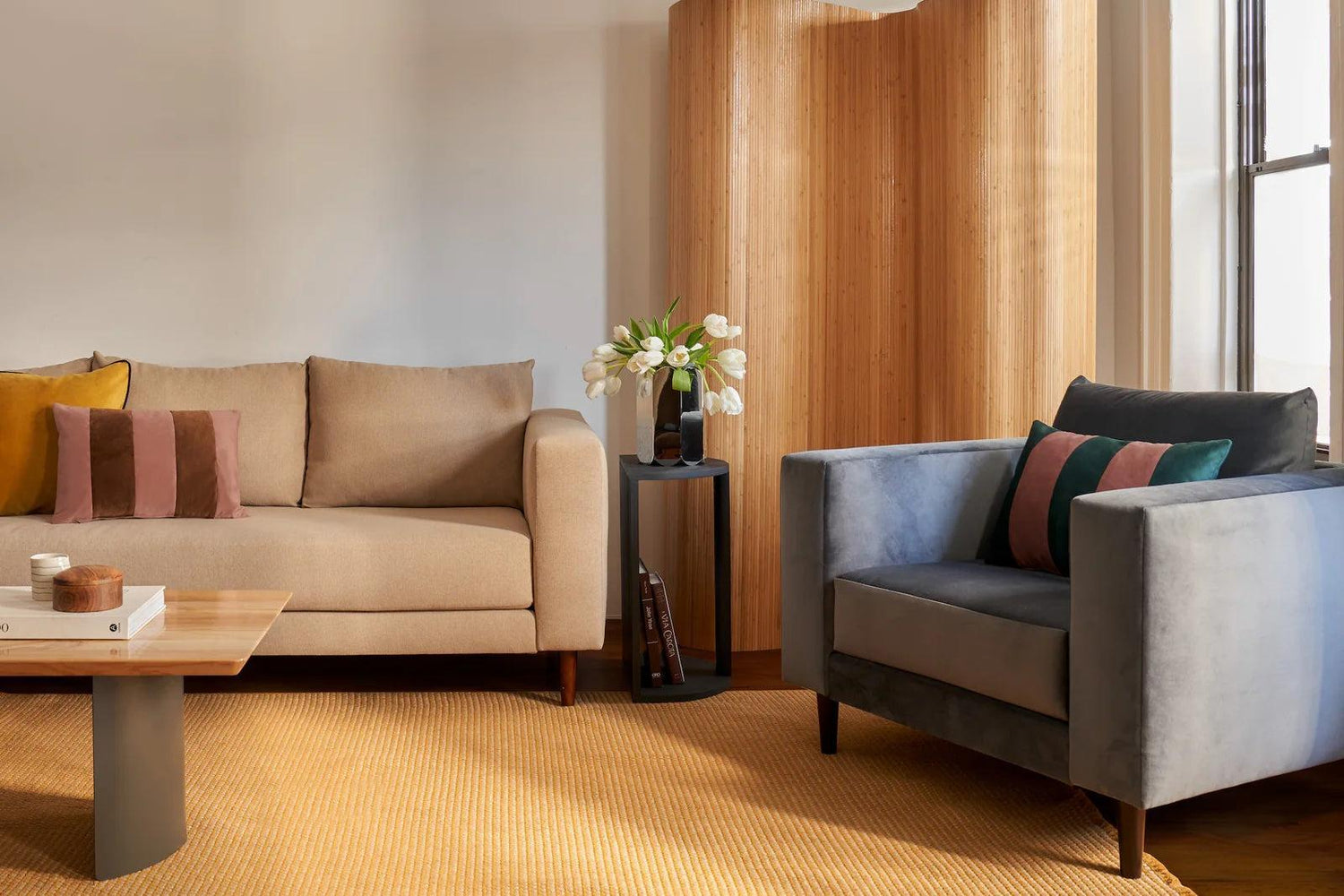 A modern living room with a beige sofa and a blue armchair, both featuring colorful throw pillows. A wooden coffee table stands on a yellow rug, and a small side table with a vase of white flowers is placed beside the sofa. A wooden room divider is in the background.