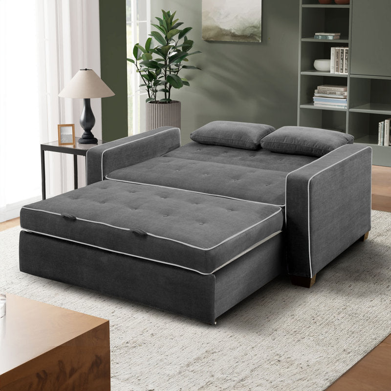 A modern living room showcases the Serta Monroe Full Size Convertible Sofa in dark gray, complemented by two cushions and set on a light-colored rug. A floor lamp and potted plant are positioned near a window adorned with sheer curtains, while a bookshelf stands in the background.