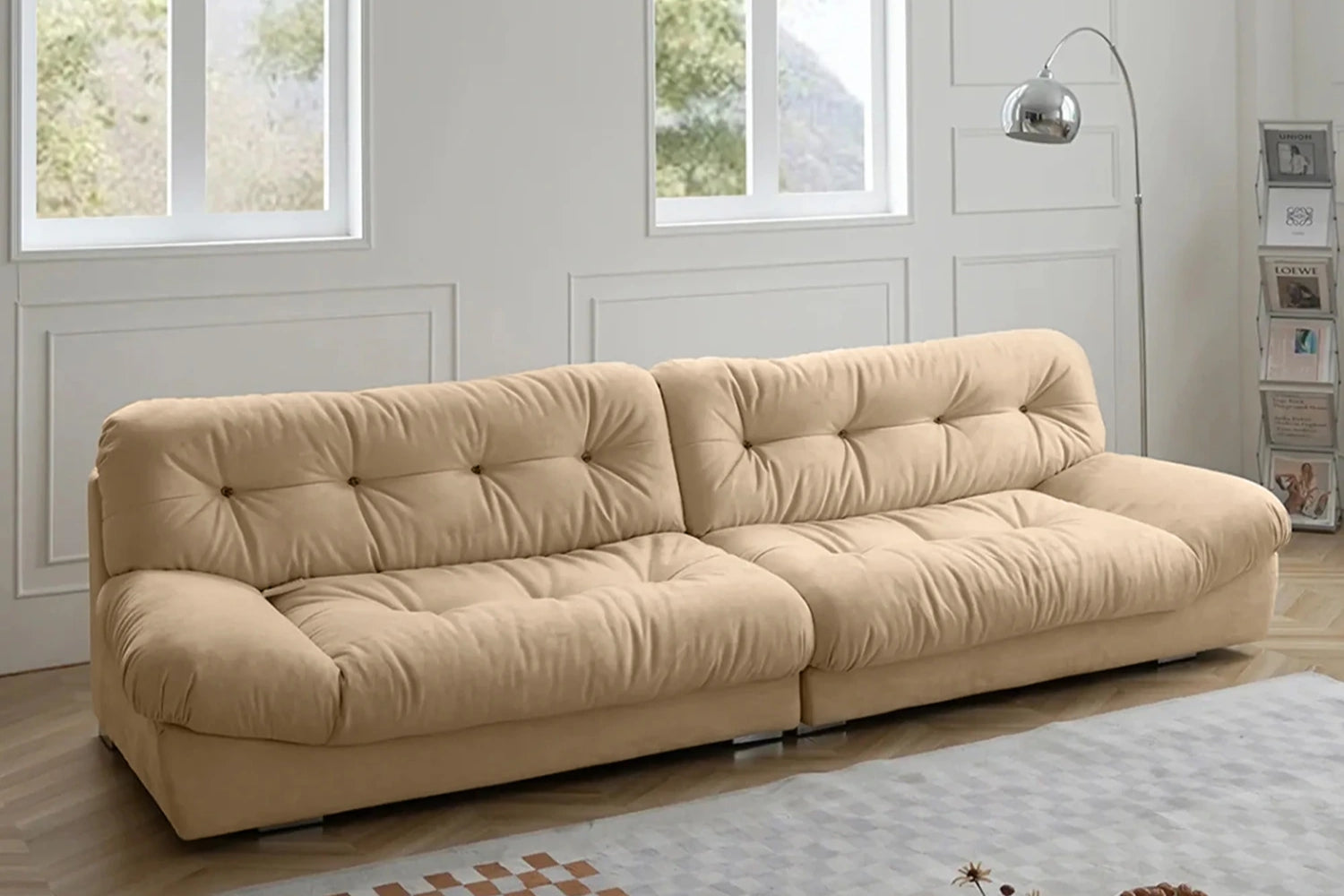 A beige, tufted sofa with a modern design sits in a living room with large windows. The sofa has two sections and is placed on a light-colored rug. A tall, arching floor lamp stands nearby, and a shelf is visible with books and decor items.