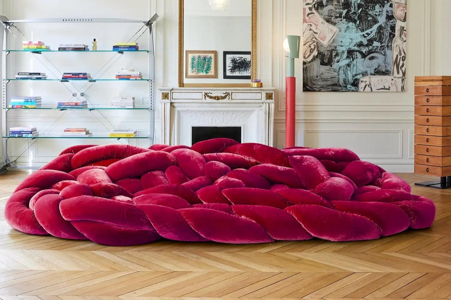 A luxurious living room with a large, braided red velvet sofa in the center. The room features a white fireplace, modern shelving with books, a tall red lamp, and framed artwork on the wall. The floor is wooden with a herringbone pattern.