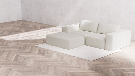 A minimalist living room features the Modular 2-Seater Chaise Sectional in Boucle Aspen complemented by a matching ottoman on a white rug. The floor showcases a herringbone wood pattern, while the plain gray walls contribute to a clean and modern aesthetic.