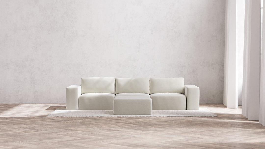 In a bright, spacious room with a light wood herringbone floor and large windows flooding the space with natural light, the Modular 3-Seater Sofa + Ottoman in Boucle Aspen sits gracefully on a textured rug. It features four square sections and is paired with a matching ottoman, creating an elegant minimalist look.