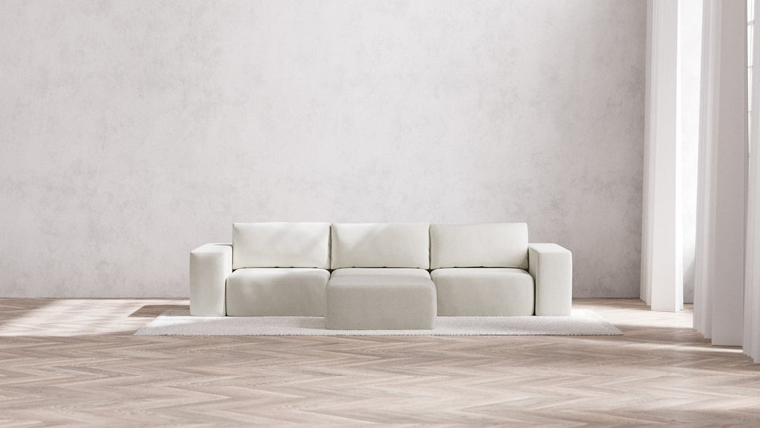 A minimalist living room showcases a Modular 3-Seater Sofa and Ottoman in Boucle Aspen, set against a light gray wall. The room is illuminated by natural light streaming through a large window, highlighting the textured wooden floor. Beneath the sofa sits a white rug.