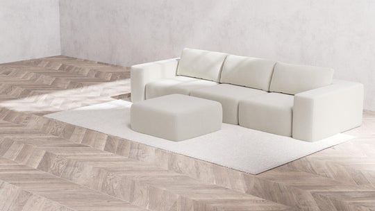 A minimalist room features a modern L-shaped Modular 3-Seater Sofa with an Ottoman in Boucle Aspen, placed on a light textured rug. The floor showcases a herringbone wood pattern, and the walls are understated with a soft grey tone.