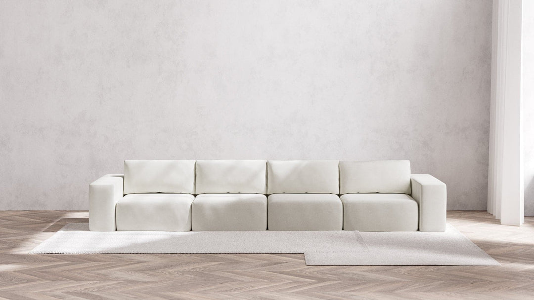 A minimalist room with a Modular 4-Seater Sofa in Boucle Aspen positioned against a light gray wall. The floor displays a herringbone wood pattern, topped with a large, light-colored rug. Natural light streams in from an unseen window.