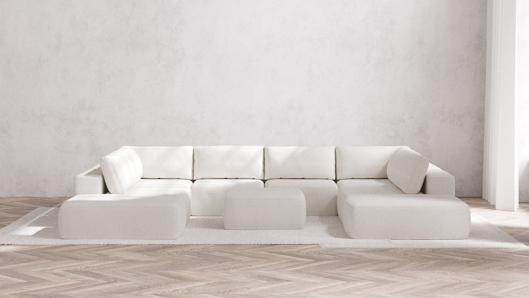 A minimalist living room features the spacious Modular 8-Seater Open-Ends U Sectional + Ottoman in Boucle Aspen, set against a light gray wall. The wooden herringbone floor brings warmth to the space, while a light area rug ties together the seating arrangement.