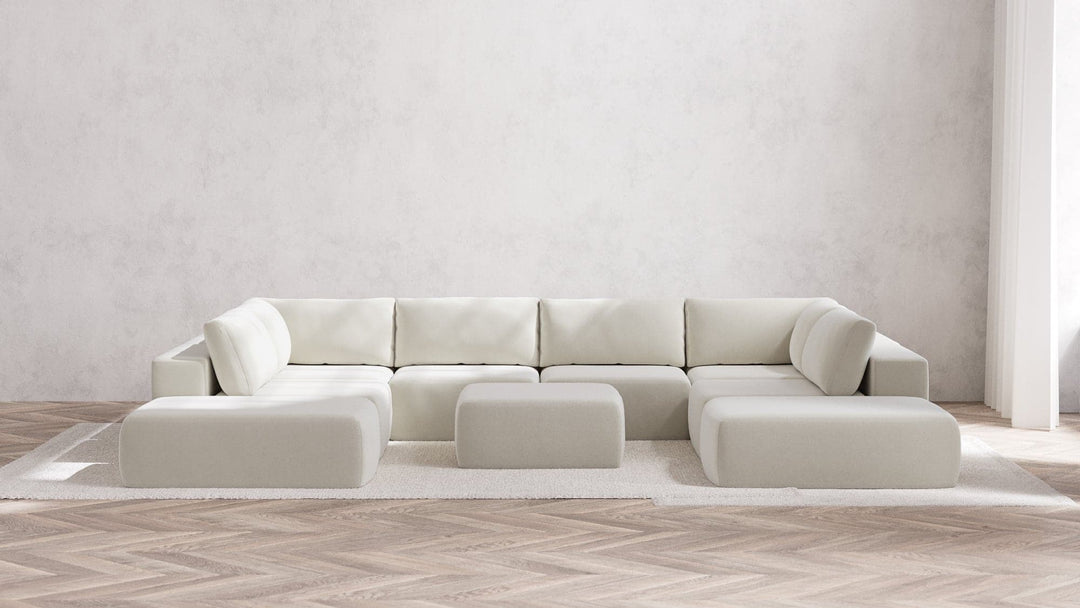 In a modern, minimalist living room, a large Modular 8-Seater Open-Ends U Sectional and matching Ottoman in Boucle Aspen take center stage on a clean white rug. The space maintains its neutral color palette, highlighted by a herringbone wooden floor and textured white walls. Soft natural light streams in from the right.
