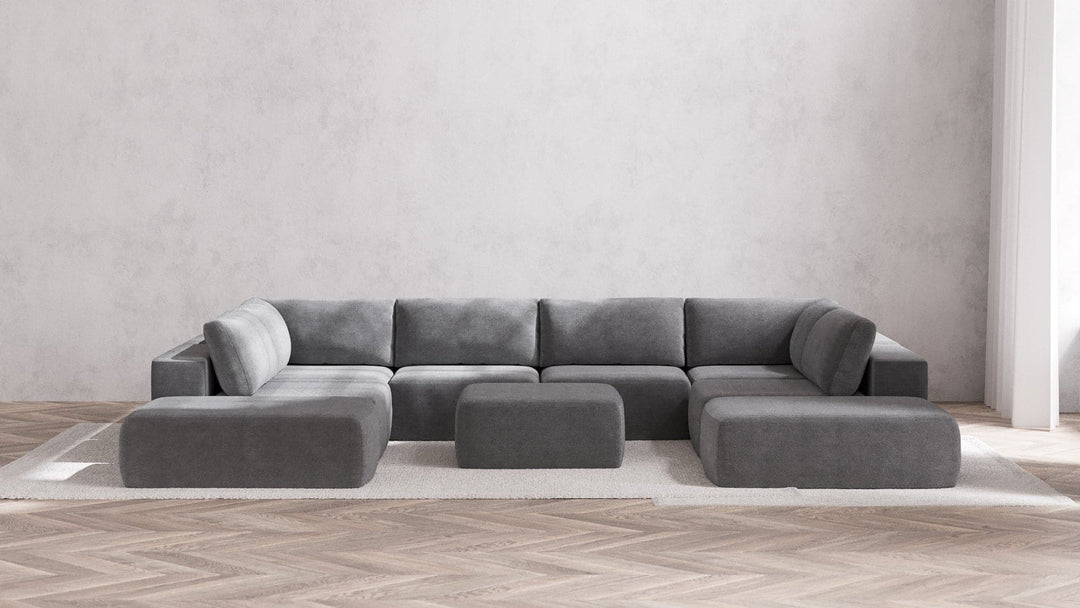 A minimalist living room is highlighted by the Modular 8-Seater Open-Ends U Sectional + Ottoman in Boucle Aspen, which boasts sleek modern lines. Two matching gray ottomans complement the arrangement, all set on a light-colored area rug atop herringbone wood flooring, framed by a plain white wall.