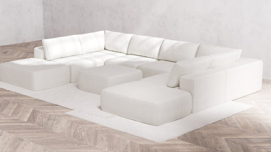 A minimalist living room showcases the Modular 8-Seater Open-Ends U Sectional + Ottoman in Boucle Aspen, providing multiple sections and cushions. The arrangement rests on a light wooden herringbone floor atop a white rug, set against a plain gray wall.