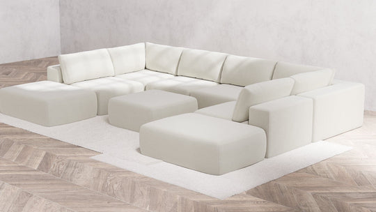 In a contemporary minimalist living room, the Aspen Boucle Modular 8-Seater Open-Ends U Sectional is prominently displayed with matching ottomans, all set on a pristine white rug. The light wood herringbone flooring and plain light gray walls enhance the clean and airy ambiance of the space.