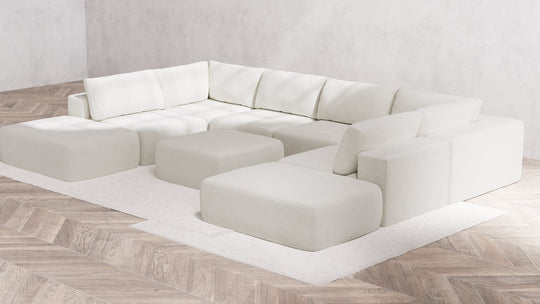 A large, contemporary white "Modular 8-Seater Open-Ends U Sectional + Ottoman | Boucle in Aspen" with firm cushions is placed in the corner of a room featuring light wood herringbone flooring. The sectional rests on a white rug, fostering a minimalist and cozy ambiance.