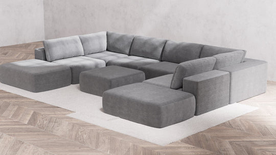 A contemporary living room showcases the Modular 8-Seater Open-Ends U Sectional with Ottoman in Boucle Aspen, elegantly positioned in a U-shape on a light-colored area rug. The space is complemented by light wood herringbone flooring and minimalist concrete walls.