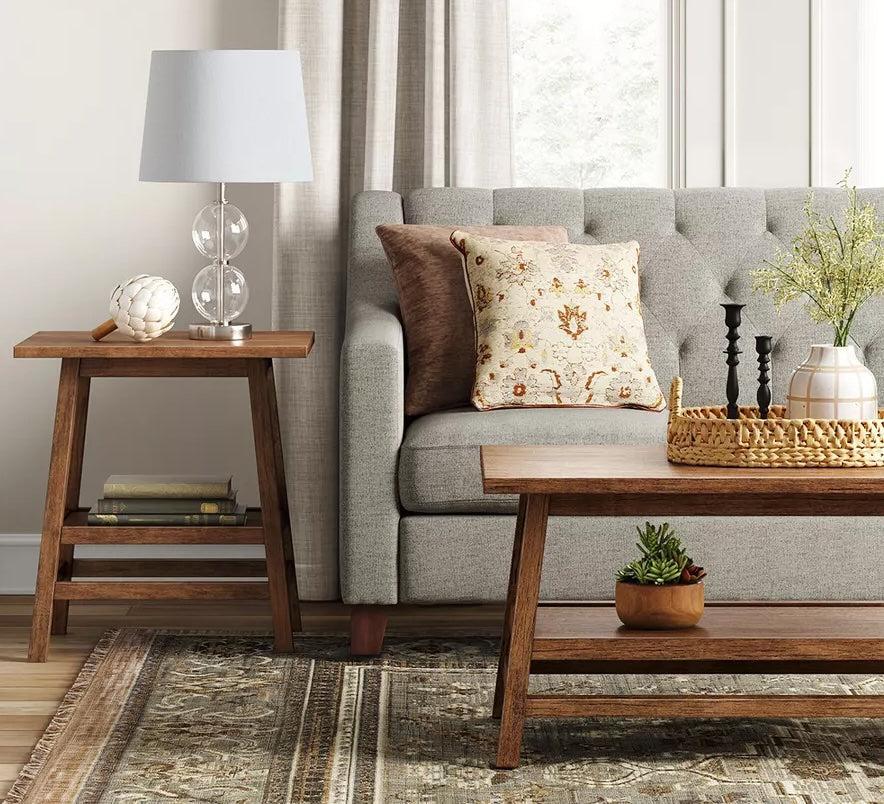 A cozy living room featuring a gray tufted sofa adorned with a patterned throw pillow. To the left is a wooden side table with a lamp and decor. In front, a wooden coffee table holds a basket, candlesticks, and a small plant. The room has a large window with curtains.