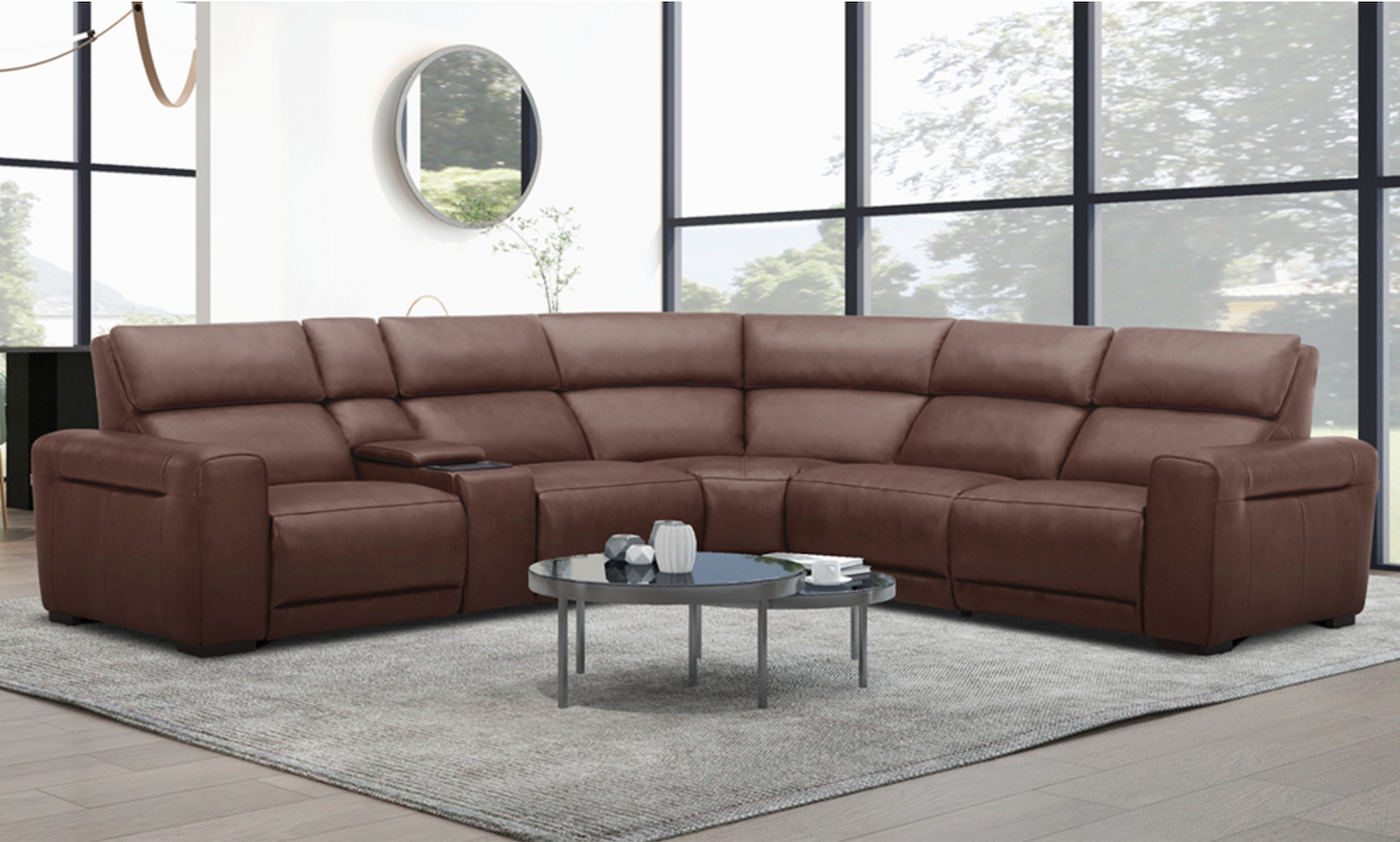 A spacious living room featuring a large, brown leather sectional sofa with a built-in console in the corner. A round coffee table sits in front of the sofa, and large windows provide a view of trees outside. A round mirror hangs on the wall.