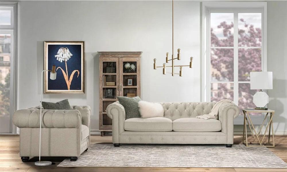 A stylish living room with a beige tufted sofa, matching armchair, and gold-accented furniture. A painting of a white flower hangs on the wall above a wooden cabinet filled with decor items. Large windows show cherry blossoms outside. A modern chandelier hangs above.