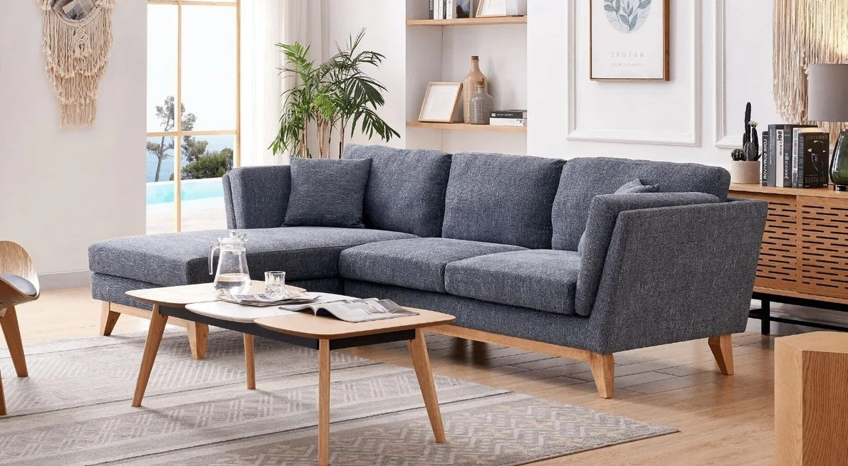 A modern living room with a gray sectional sofa, a wooden coffee table with a black top, and various decorative items. There's a potted plant by the window, wall art, and shelves with books and decor. A large window provides natural light and a view outside.