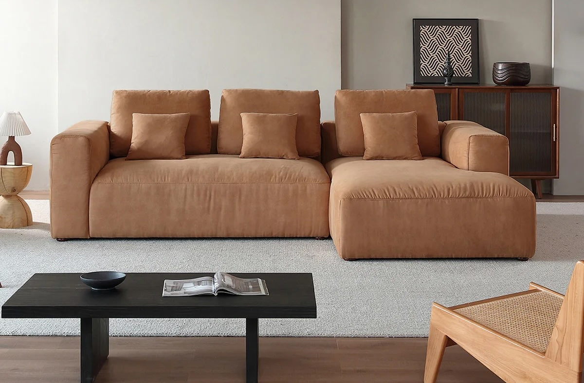 A modern living room featuring a large, tan sectional sofa with several matching cushions. A black coffee table with an open book and bowl sits in front of the sofa. In the background, there is a wooden cabinet with decorative items and a light beige wall.