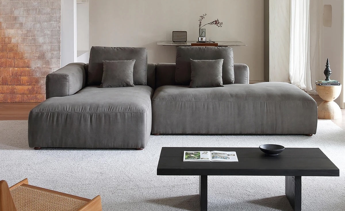 A modern living room features a large gray sectional sofa adorned with matching cushions. A black coffee table with a magazine and a bowl sits on a light-colored carpet. In the background, there's minimalistic decor with a small plant and items on a shelf.