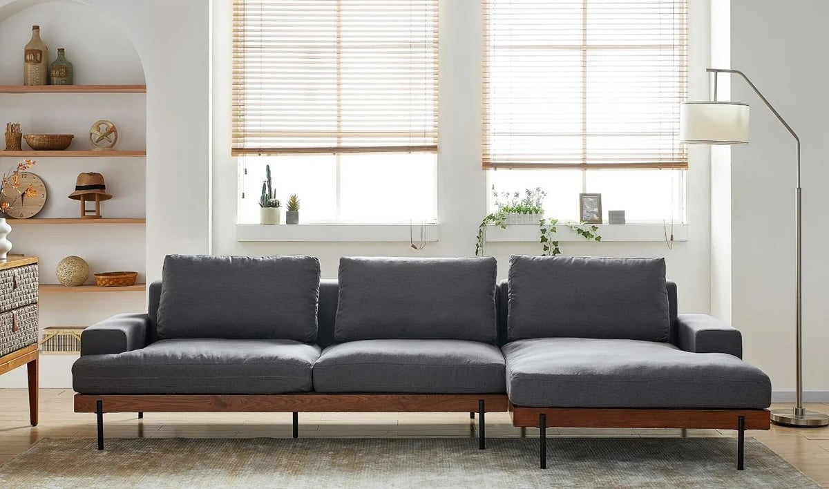 A modern living room features a large gray sectional sofa with wooden legs, flanked by two windows with light wooden blinds. The room also includes a floor lamp, potted plants on the windowsill, and a set of shelves with decorative items on the left.