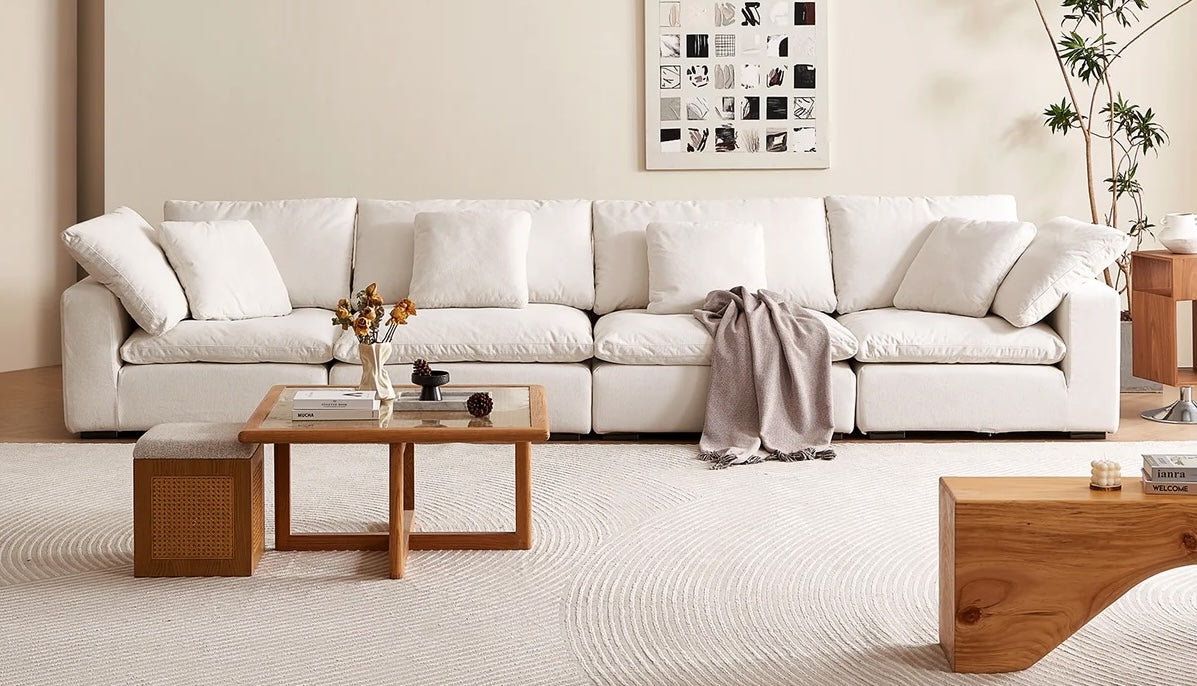 A spacious, minimalist living room features a long white sectional sofa adorned with white cushions and a draped gray blanket. A wooden coffee table with decor items is in front of the sofa, and wall art and a potted plant add to the serene ambiance.