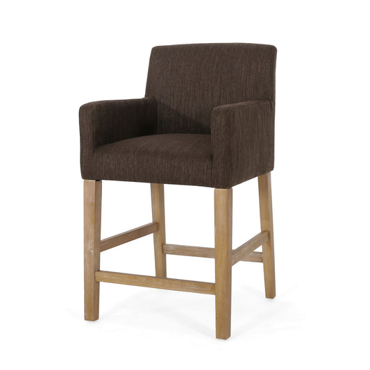 The Charmed Upholstered Counter Stool features a brown upholstered seat, backrest, and round arms. It has light wood legs with support bars in an H-frame base for added stability.