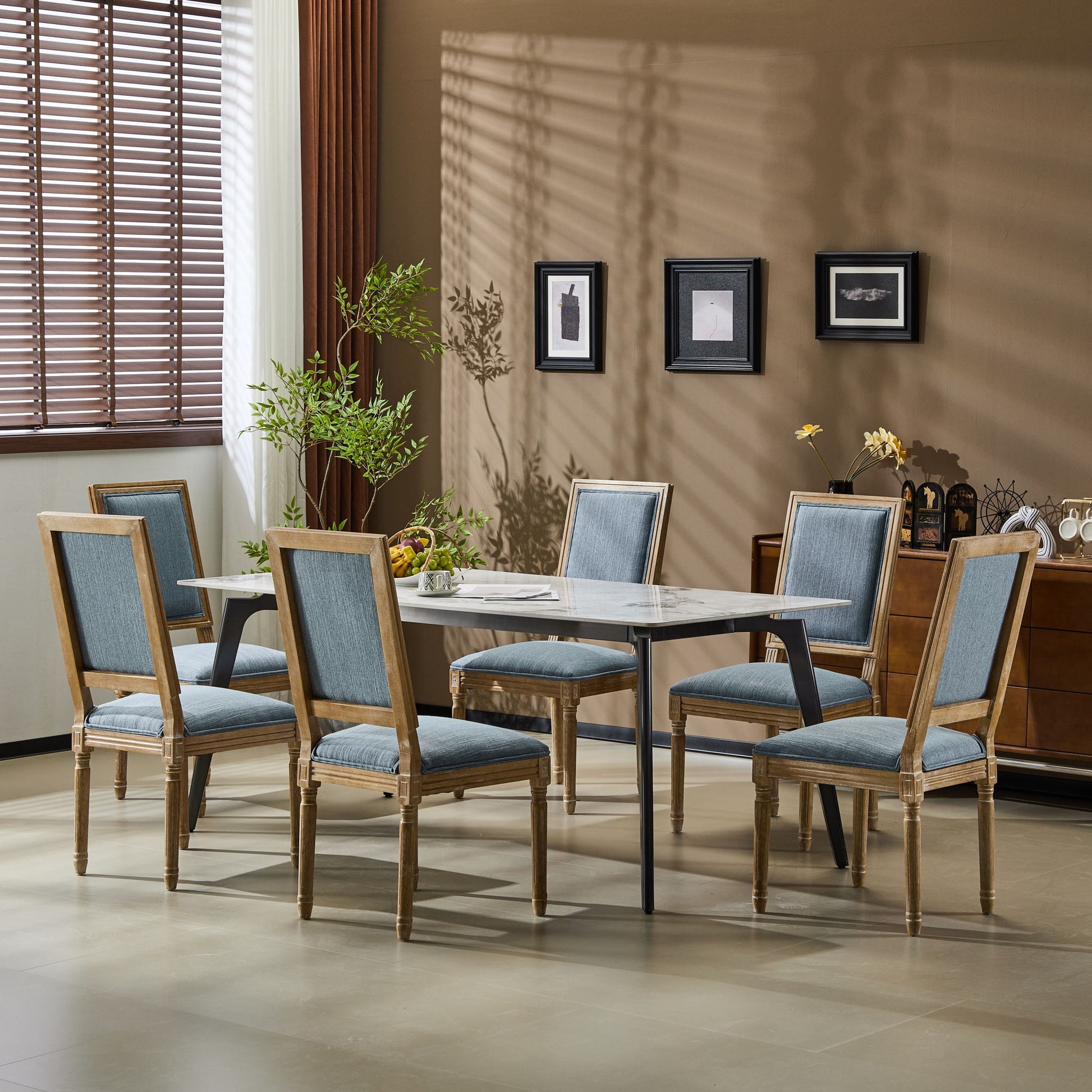 A modern dining room features a rectangular table with six French Country Style Dining Chairs with upholstered seating and fluted legs, a potted plant, wall art, a sideboard under window blinds, and sunlight casting shadows on the beige wall.
