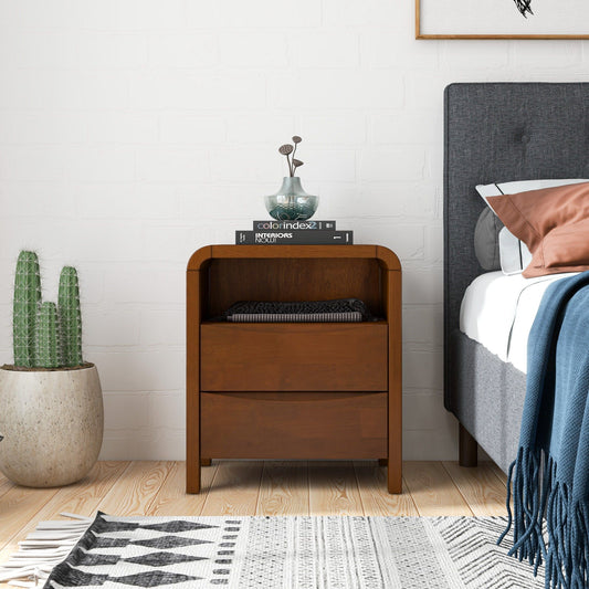 The Lionel Mid-Century Modern Nightstand 2 Drawer in brown sits by the bed, topped with books, a glass bowl, and a decorative sculpture. A potted cactus and a patterned rug are placed on the floor next to it.