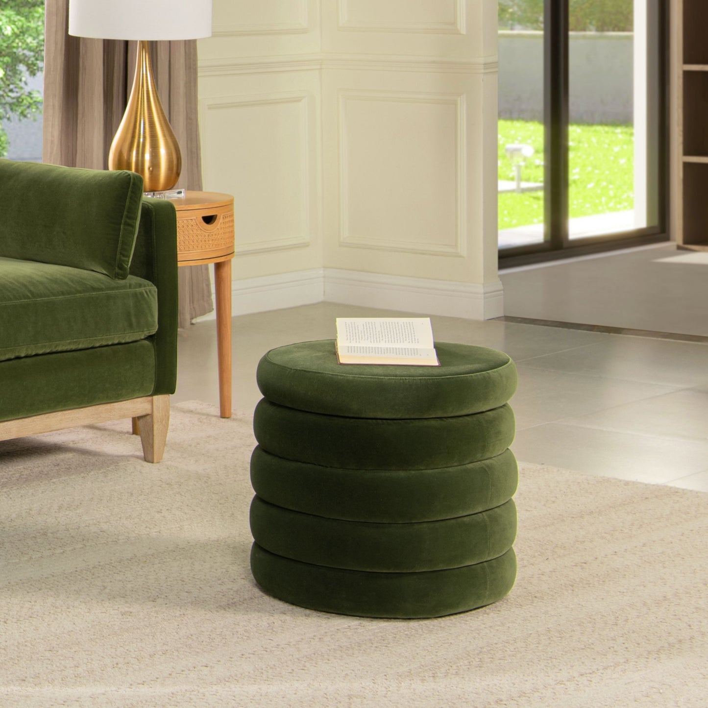 The Fuji Oversized Round Storage Ottoman, upholstered in green velvet with a stacked ring design, sits on a beige rug beside a matching sofa. An open book rests on top as sunlight pours through large glass doors in the modern living room.