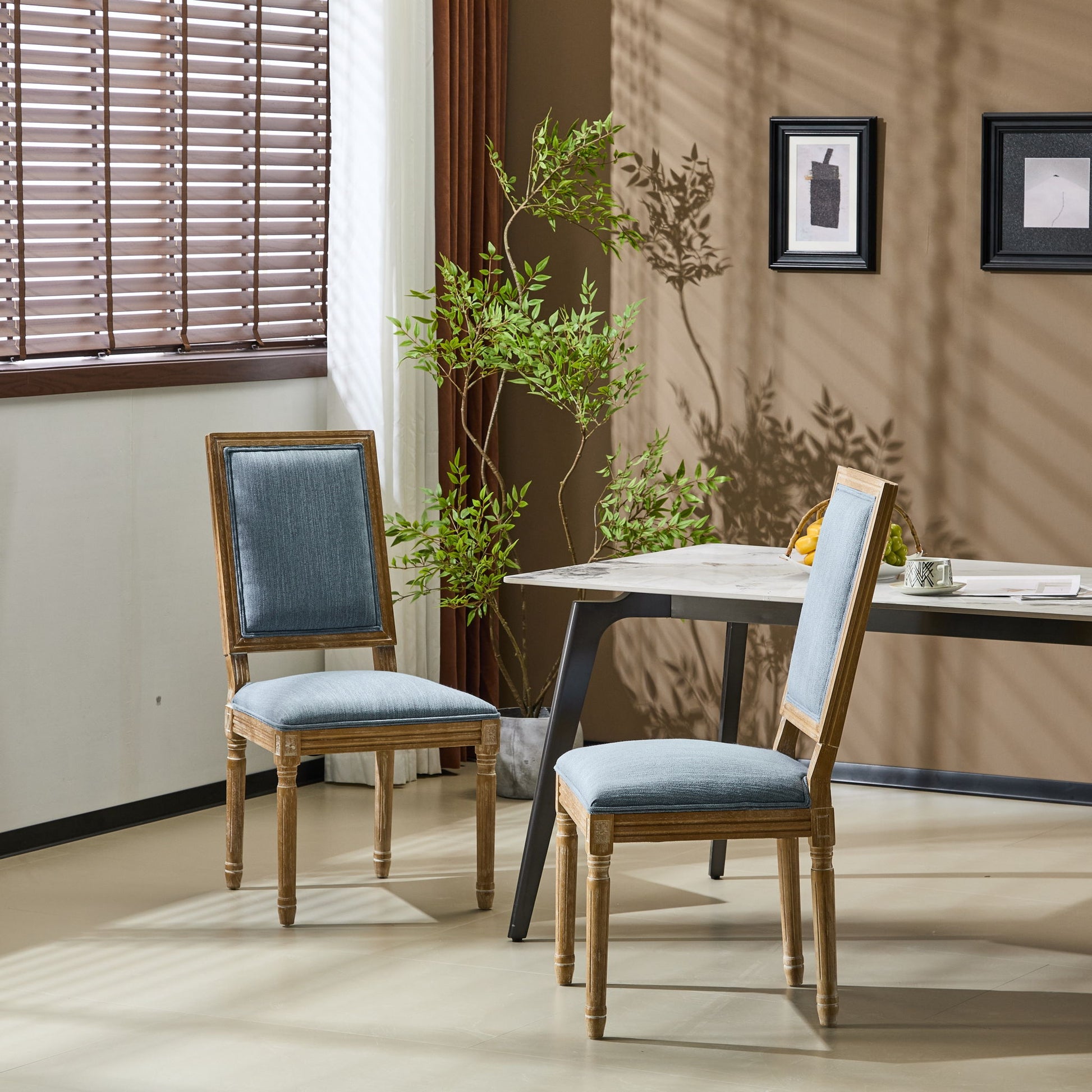 Two French Country Style Dining Chairs with upholstered seating and fluted legs, featuring light blue cushions, sit by a modern black table in a sunlit room with beige walls, plants, framed art, and a window with brown blinds.